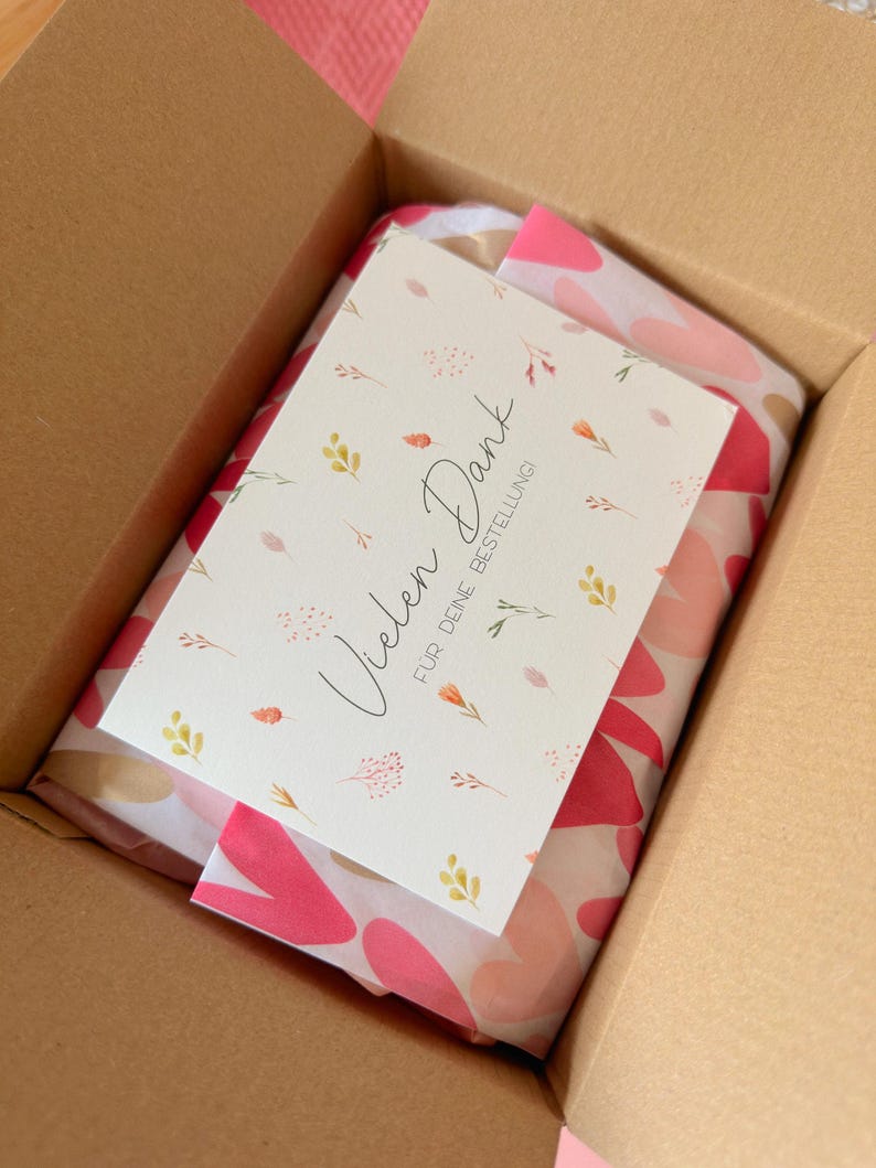 May include: A brown cardboard shipping box containing a package wrapped in white tissue paper with a pink and red heart pattern. A white rectangular card rests on top, featuring small floral illustrations and the text Vielen Dank F&Uuml;R DEINE BESTELLUNG.