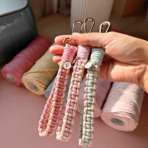 Pode incluir: Três porta-chaves de pulso em macramé com cordão entrançado em rosa, lavanda e azul claro, cada um combinado com um cordão creme. Cada porta-chaves inclui um mosquetão de metal dourado e um pequeno pendente circular com uma letra.