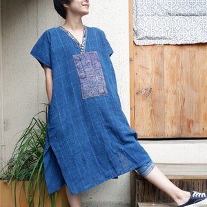 May include: A blue knee-length tunic dress featuring a subtle grid pattern, a V-neckline with decorative trim, and a rectangular embroidered panel on the chest with intricate geometric patterns in red, white, and blue thread accents.