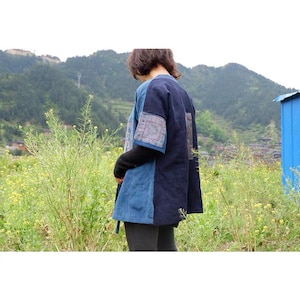 May include: A short-sleeved jacket featuring a color-blocked design with a light blue front panel and a dark navy blue back panel. The sleeves and back are accented with rectangular patches displaying intricate geometric patterns in muted tones.
