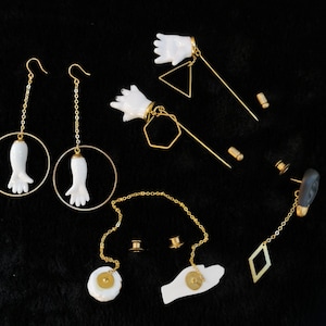 May include: A collection of unique jewelry pieces featuring white hand-shaped pendants and charms. The earrings and pins are accented with gold-colored chains, hoops, and geometric shapes. The jewelry is displayed on a black surface.