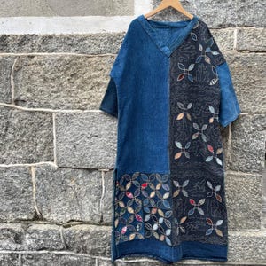May include: A knee-length dress featuring a V-neckline and short sleeves. The garment is constructed from a combination of solid blue denim fabric and a contrasting black panel adorned with intricate white stitched patterns and colorful floral motifs.