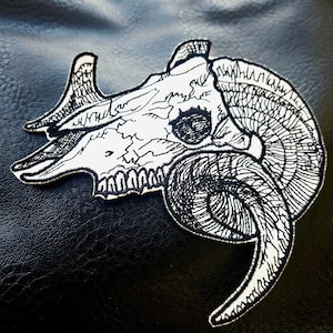 May include: A white embroidered patch depicting a ram skull with detailed horns. The skull is outlined in black thread and the horns are intricately detailed.