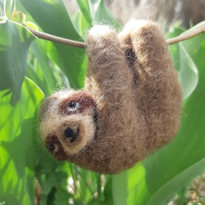 May include: A brown and white needle felted sloth hanging upside down from a branch. The sloth has a cute face with black eyes and a small black nose.