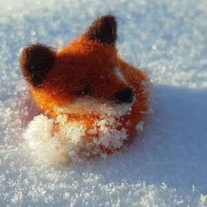 May include: A small orange and brown felt fox toy is lying in the snow. The fox has a fluffy tail and a cute face.