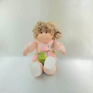 May include: A vintage doll with curly blonde hair, wearing a pink and green patterned outfit with white trim. The doll has a soft, plush body and large, white shoes. The doll is sitting with its arms outstretched.