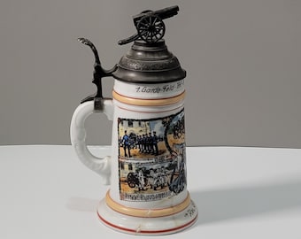 May include: A white ceramic beer stein with a pewter lid and a cannon on top. The stein features a colorful image of a group of soldiers and a cannon. The text on the stein reads "7. Garde-Feld-Art. Erinnerungs-Anstalt."