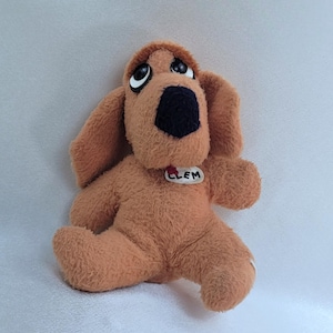 May include: A plush brown dog toy with large, expressive eyes and a black nose. The dog has floppy ears and a small tag that reads "CLEM". The toy is sitting on a white surface.
