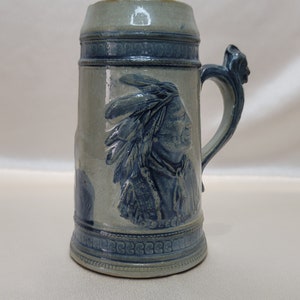 May include: A blue and white ceramic beer stein with a relief image of an indigenous person wearing a headdress. The stein has a handle and the words "As Sleep" are visible on the front.