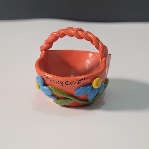 May include: A small orange ceramic basket with a twisted rope handle and blue and yellow flowers painted on the side. The basket says "Fairycave" on the side.