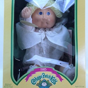 May include: A vintage Cabbage Patch Kids doll in its original packaging. The doll has blonde yarn hair, blue eyes, and is wearing a white dress and sweater. The box features the Cabbage Patch Kids logo, the year 1985, and the phrase "I'm unique. I'm adoptable!"