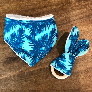 May include: A baby bib and a bunny ear teether set. Both items feature a blue and turquoise tropical leaf pattern with white trim. The teether has a wooden ring.