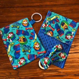 May include: A blue and green baby blanket with a Paw Patrol theme. The blanket features a variety of characters from the show, including Chase, Marshall, and Skye. The blanket also includes a small, blue and green teether with a wooden ring.