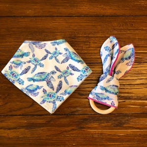 May include: A white baby bib and a matching teether with a blue and purple watercolor sea turtle pattern. The teether has a wooden ring.