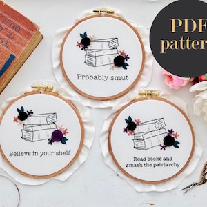 May include: Three embroidery hoop art pieces with a stack of books and floral designs. The text on the hoops reads "Probably smut", "Believe in your shelf", and "Read books and smash the patriarchy".