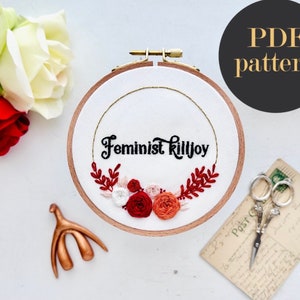 May include: Embroidered hoop art featuring the text "Feminist Killjoy" in black cursive lettering. The hoop is decorated with red, white and orange flowers and green leaves.