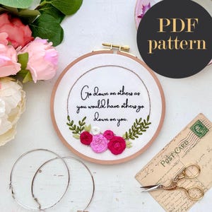 Puede incluir: Arte de bordado en aro con la cita: "Go down on others as you would have others go down on you." El aro está decorado con flores rosas y blancas.