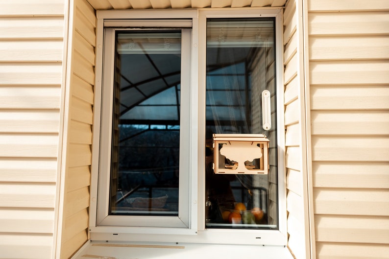 Peut inclure: Un mangeoire &agrave; oiseaux en bois avec un motif d'arbre est fix&eacute;e &agrave; l'ext&eacute;rieur d'une fen&ecirc;tre. La mangeoire est en bois brun clair et a un fond en plastique transparent. La fen&ecirc;tre est blanche et a un vitrage coulissant. La mangeoire est suspendue au sommet du cadre de la fen&ecirc;tre.