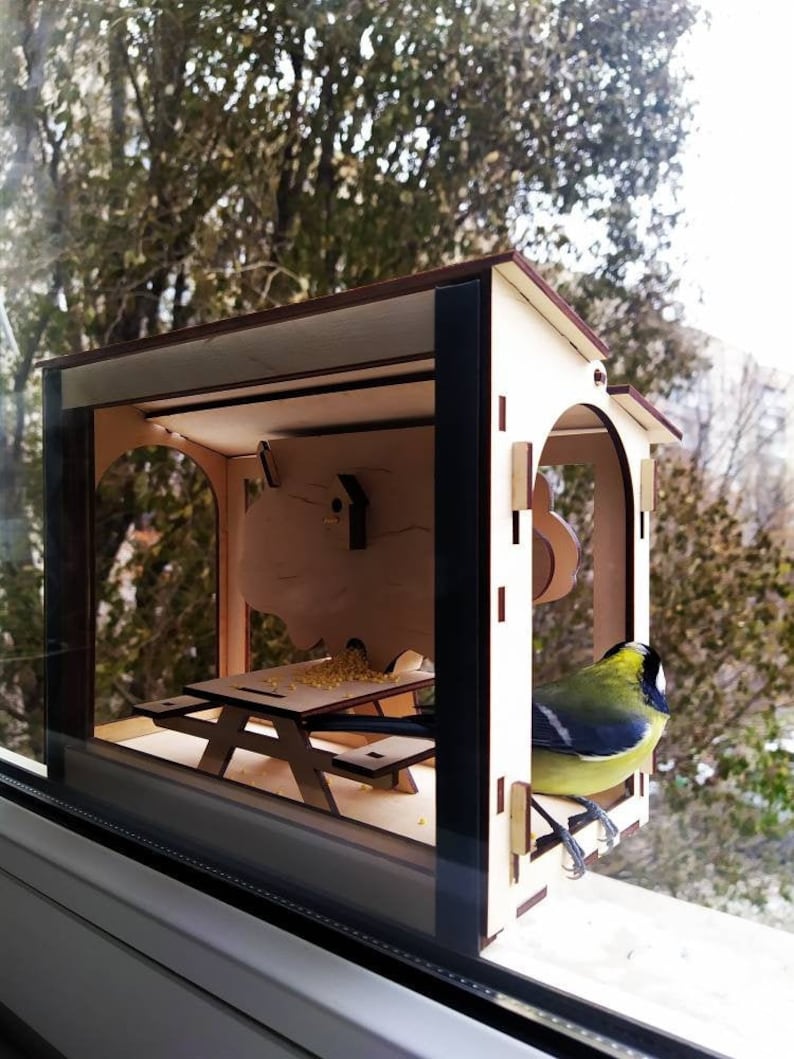 Peut inclure: Un mangeoire &agrave; oiseaux en bois avec une petite table de pique-nique &agrave; l'int&eacute;rieur. Un oiseau jaune et bleu est perch&eacute; sur le bord de la mangeoire.