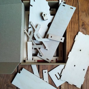 Peut inclure: Une bo&icirc;te en carton avec le couvercle ouvert, r&eacute;v&eacute;lant un ensemble de pi&egrave;ces en bois pour un projet d'artisanat. Les pi&egrave;ces sont de formes et de tailles vari&eacute;es, et certaines ont de petits trous pour l'assemblage. La bo&icirc;te est pos&eacute;e sur une surface en bois.