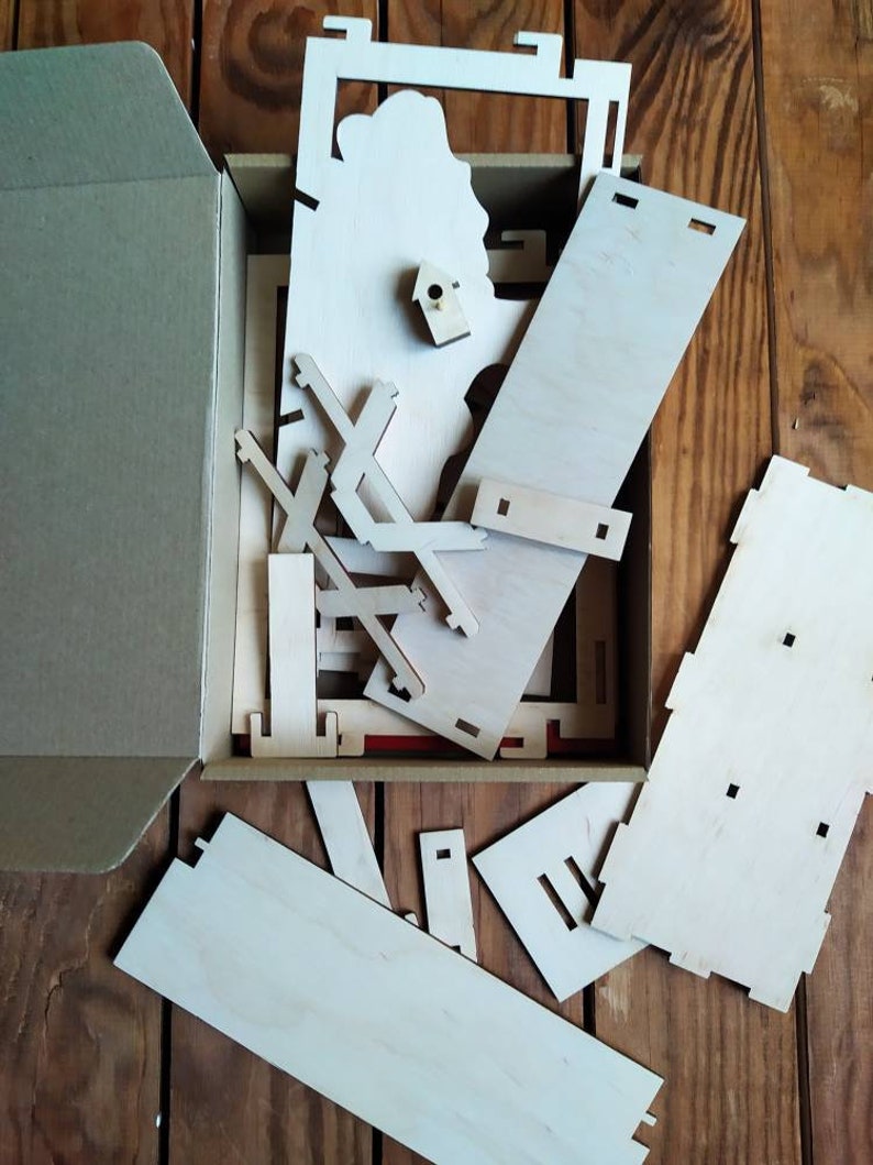 Peut inclure: Une bo&icirc;te en carton avec le couvercle ouvert, r&eacute;v&eacute;lant un ensemble de pi&egrave;ces en bois pour un projet d'artisanat. Les pi&egrave;ces sont de formes et de tailles vari&eacute;es, et certaines ont de petits trous pour l'assemblage. La bo&icirc;te est pos&eacute;e sur une surface en bois.