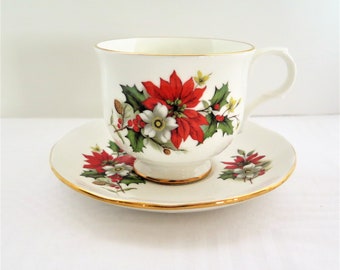 May include: A white teacup and saucer with a gold rim. The cup and saucer feature a floral design with red poinsettias, white flowers, green leaves, and red berries.