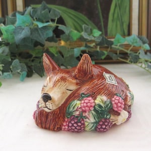 May include: A ceramic fox figurine in shades of brown, cream, and pink, asleep amongst raspberries and green leaves. The fox is curled up, with a small tag attached. A decorative item for a home.