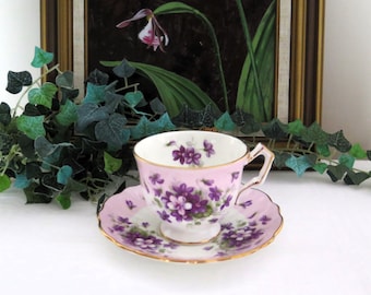 Può includere: Una tazzina da tè e un piattino rosa con un delicato motivo floreale con violette viola. La tazza e il piattino hanno un bordo dorato.