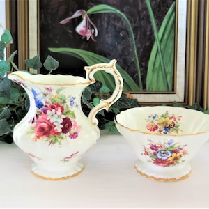 Peut inclure: Un ensemble de crémier et de sucrier en porcelaine blanche avec un motif floral dans des tons de rose, rouge, jaune et bleu. L'ensemble est orné de bordures dorées et d'un bord festonné.
