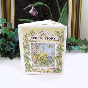 May include: A hardback book titled "Spring Story" by Jill Barklem. The cover displays a colourful illustration of yellow flowers and a woodland scene, framed by green vines and the book's title. The book is open, showing white pages.