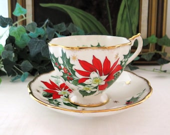 Könnte beinhalten: Ein weißes Porzellan-Teeservice bestehend aus Tasse und Untertasse mit einem weihnachtlichen Motiv aus roten Weihnachtssternen, grünen Stechpalmenblättern und kleinen roten Beeren. Die gewellten Ränder sind mit einem dünnen Goldrand verziert.