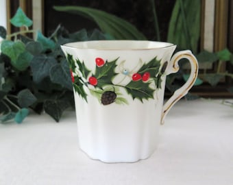 Könnte beinhalten: Eine weiße Porzellantasse mit goldfarbenem Rand und Henkel. Die Tasse zeigt ein festliches Design mit Stechpalmenblättern, roten Beeren und Tannenzapfen.