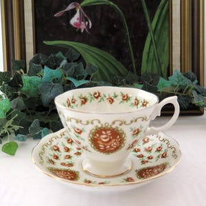 Può includere: Servizio da tè color crema con tazza e piattino. Il motivo floreale presenta piccole rose rosse e bordi dorati. Un medaglione floreale più grande è centrato sulla tazza e sul piattino.