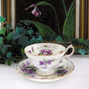 May include: A white teacup and saucer with a delicate floral pattern of purple violets. The cup has a gold rim and a handle. The saucer has a matching floral pattern and a gold rim.