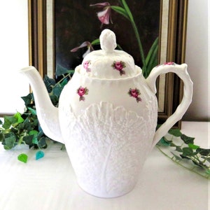 Może przedstawiać: Biały porcelanowy czajnik z wzorem liścia kapusty i różowymi akcentami róż. Czajnik ma pokrywkę z gałką w kształcie pąka róży.