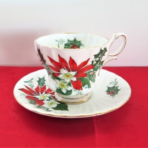 May include: A white teacup and saucer with a gold trim. The cup and saucer are decorated with a red poinsettia flower, green holly leaves, and white flowers.