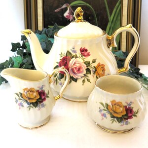 May include: A vintage cream porcelain tea set, comprising a teapot, creamer jug, and sugar bowl. Each piece is decorated with a floral pattern of red, pink, and yellow roses. The teapot and creamer have gold detailing.