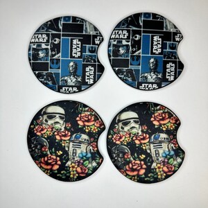May include: Set of four round car coasters featuring Star Wars characters. Two coasters feature a collage of Star Wars characters and the text "Star Wars" in a blue and black pattern. The other two coasters feature a black background with floral designs and the characters R2-D2 and a Stormtrooper.