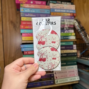 May include: Two white car coasters with a red cherry and bow pattern. The coasters are packaged in a clear plastic sleeve with the text "Car Coasters" printed on the front.