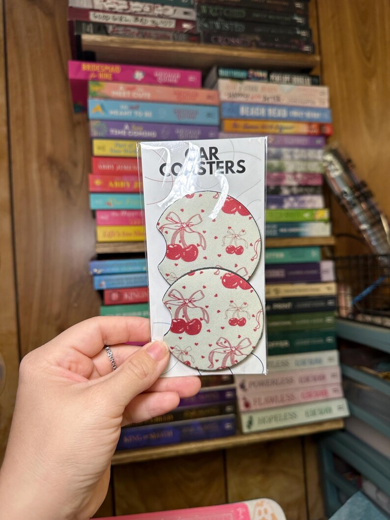 May include: Two white car coasters with a red cherry and bow pattern. The coasters are packaged in a clear plastic sleeve with the text "Car Coasters" printed on the front.