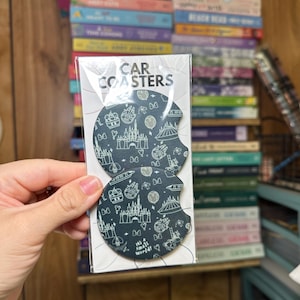 May include: Two black round car coasters with a white outline of various Disney attractions, including Cinderella's Castle, Spaceship Earth, and the text "It's a small world".