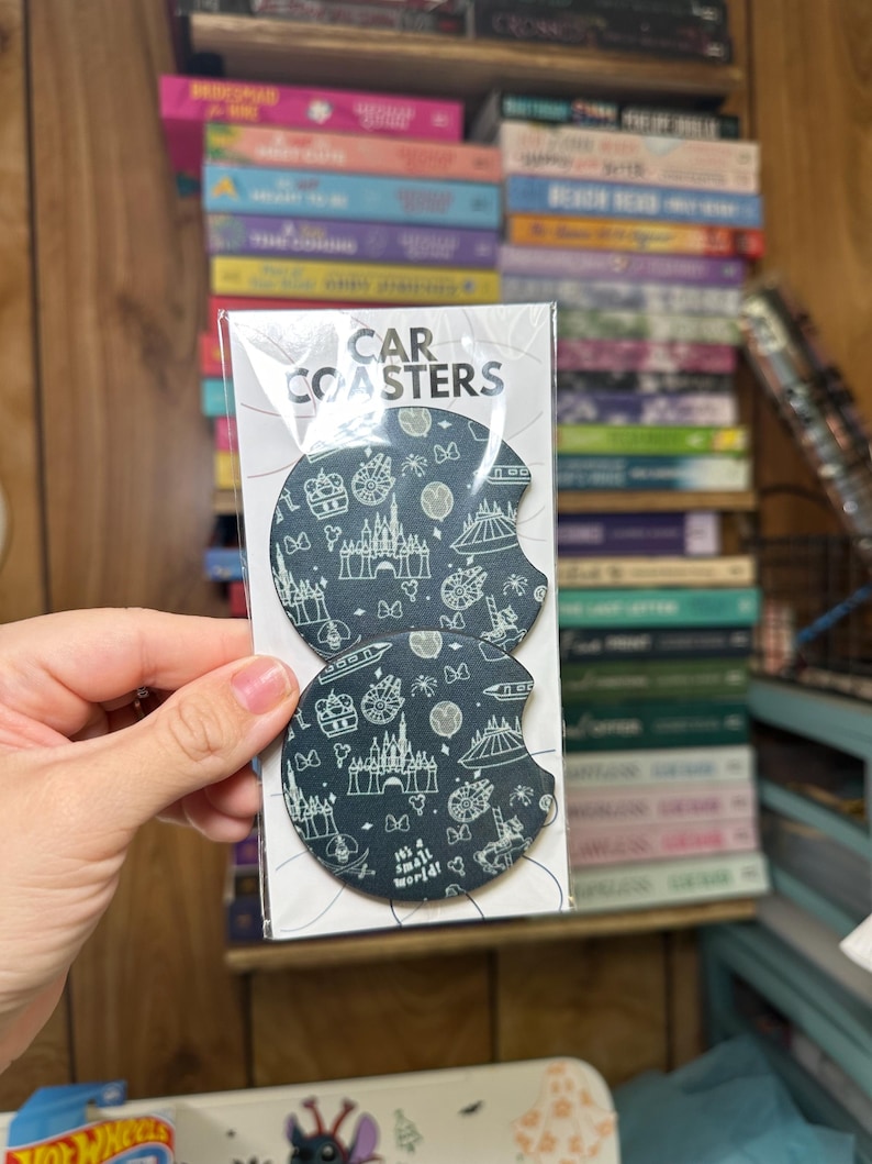 May include: Two black round car coasters with a white outline of various Disney attractions, including Cinderella's Castle, Spaceship Earth, and the text "It's a small world".