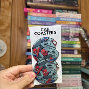 May include: Two round car coasters featuring a Spiderman design. The coasters are gray with red and black accents. The text "CAR COASTERS" is printed on the white packaging.