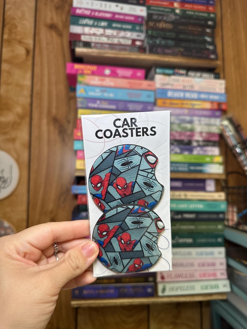 May include: Two round car coasters featuring a Spiderman design. The coasters are gray with red and black accents. The text "CAR COASTERS" is printed on the white packaging.