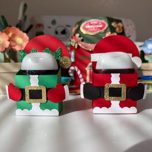 Puede incluir: Dos soportes decorativos con temática navideña, uno verde y otro rojo. El soporte verde está diseñado como un elfo, con un gorro rojo, cuerpo verde y guantes rojos. El soporte rojo está diseñado como Papá Noel, con gorro rojo, cuerpo rojo y botas negras.