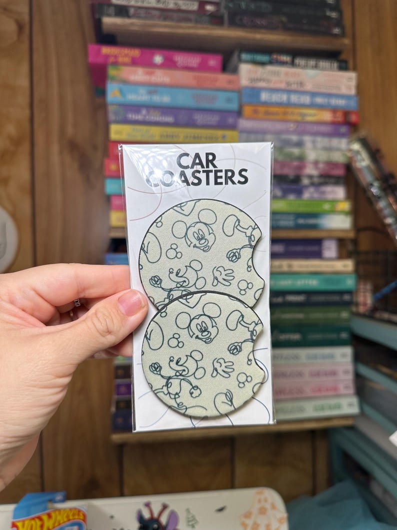 May include: Two round car coasters with a white background and a black outline of Mickey Mouse. The coasters have the text "CAR COASTERS" printed on the packaging.
