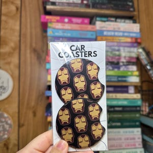 May include: Two round black car coasters with a repeating pattern of gold Iron Man masks. The packaging says "CAR COASTERS".