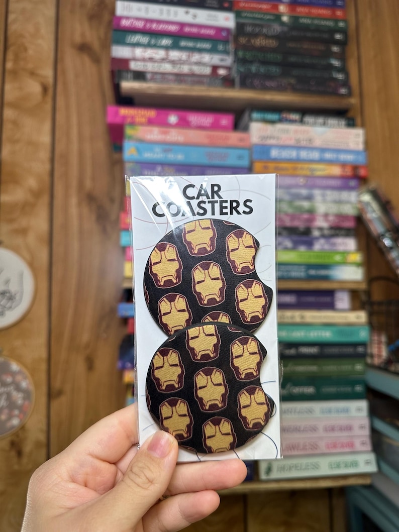 May include: Two round black car coasters with a repeating pattern of gold Iron Man masks. The packaging says "CAR COASTERS".