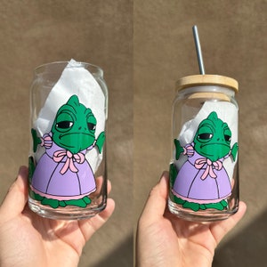 Puede incluir: Dos vasos de vidrio transparente con una pajita. Cada vaso está decorado con un camaleón de dibujos animados que lleva un vestido morado y un lazo rosa. El camaleón es verde con la barriga verde claro y un ojo azul claro.