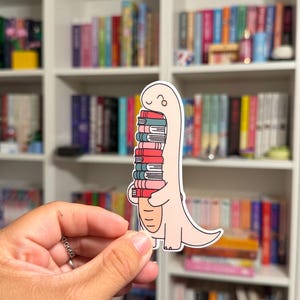 May include: A die-cut sticker featuring a light pink cartoon dinosaur with a long neck, holding a vertical stack of six colorful books in shades of red, teal, and pink. The dinosaur has a simple line-drawn face and a rounded body shape.